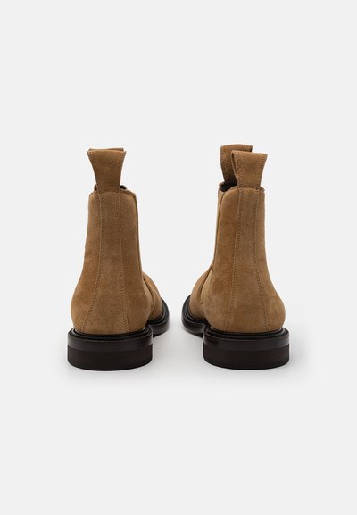 Botas Chelsea de ante marrón con suela de cuero lisa, que cuentan con una lengua en el talón para facilitar el calzado y detalles de costura en la parte posterior.