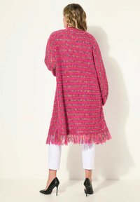 Długi, otwarty kardigan szydełkowy w intensywnym różu z wielokolorowymi wzorami, z długimi rękawami i frędzlami na dolnym końcu. Zestawiony z białymi spodniami i obcasami.