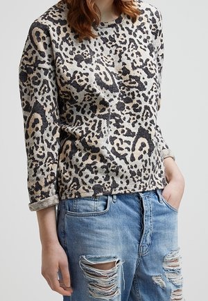 Femme portant une chemise à manches longues imprimé léopard et un jean bleu clair déchiré, une main dans la poche, sur un fond uni.