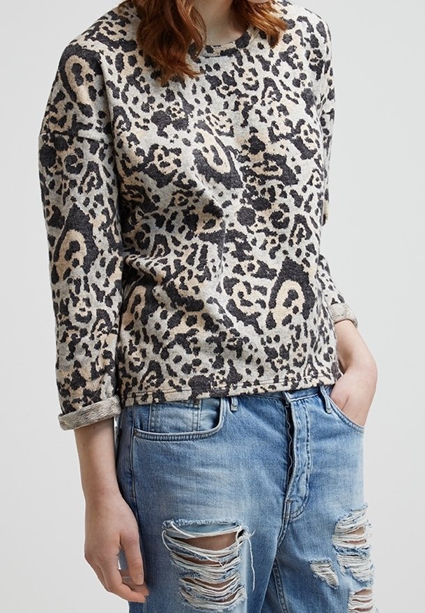Femme portant une chemise à manches longues imprimé léopard et un jean bleu clair déchiré, une main dans la poche, sur un fond uni.