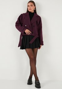 Vijolična dvoredna jakna iz teksturirane tkanine z pasom, v kombinaciji z črno plisirano krilo in polprosojnimi nogavicami.