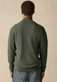Grüner, texturierter Pullover mit Kragen, gerippten Bündchen und Saum, kombiniert mit blauen Jeans. Ansicht von hinten.