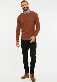 Brauner Strickpullover mit strukturierten oberen Paneelen, kombiniert mit einem karierten Hemd, schwarzen schmal geschnittenen Jeans und braunen Lederstiefeln.