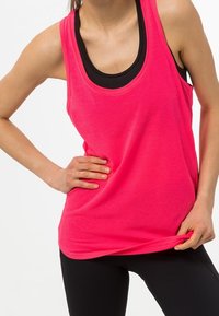 Světle růžová bezrukávová tílko s kulatým výstřihem, vyrobené z texturované látky. Nosené přes černý těsný top, v kombinaci s černými legínami.