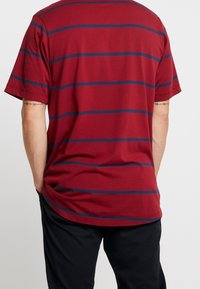 T-shirt bordeaux avec des rayures horizontales bleu foncé ; manches courtes et coupe ample ; tissu en coton doux ; sans logo ni motif visible.
