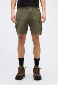 Άνδρας που φοράει cargo σορτς ελιάς, μαύρες κάλτσες μέχρι τη μέση της γάμπας και καφέ παπούτσια πεζοπορίας, στέκεται μπροστά σε απλό ανοιχτόχρωμο φόντο.