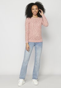 Šviesiai rožinė tekstūruota ilgomis rankovėmis megztinis derinamas su šviesiai mėlynomis džinsomis ir baltais sportbačiais. Fonas minimalus, šviesiai pilkos spalvos.