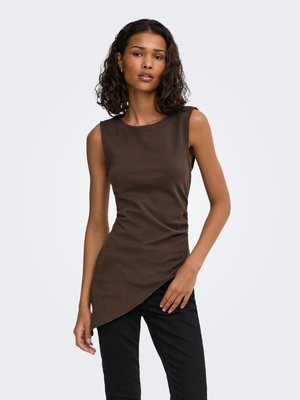 Femme aux cheveux bouclés et foncés portant un haut asymétrique sans manches marron foncé et un pantalon noir, debout devant un fond clair uni.