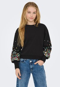 Svart sweatshirt med blommig broderi på ärmarna, ribbad halsringning och mudd, tillsammans med blå denimjeans. Mjuk tygkänsla.