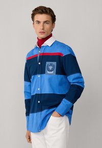 Overhemd met knoopsluiting en horizontale strepen in lichtblauw, donkerblauw en rood. Bevat een borstzak met een embleem, een witte kraag en lange mouwen.