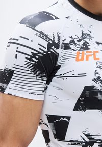 Αθλητής που φοράει στενή λευκή και μαύρη μπλούζα UFC με αφηρημένο μοτίβο και πορτοκαλί λογότυπο UFC στο στήθος.