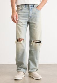 Jeans de mezclilla azul claro desgastados con grandes aberturas deshilachadas en las rodillas, un ajuste relajado y un acabado desvanecido, combinados con zapatillas beige.