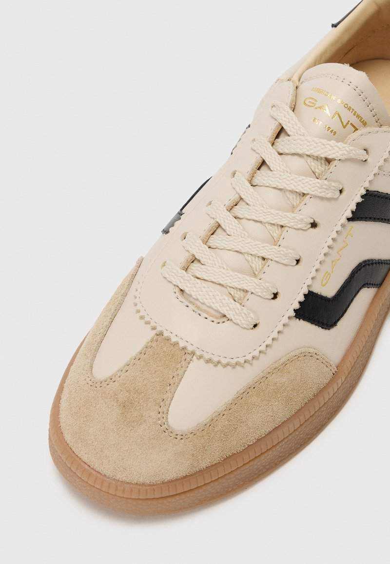 Beige Leder-Sneaker mit schwarzen Streifen, Wildlederakzenten und strukturierten Details. Ausgestattet mit runden Schnürsenkeln und einer Gummisohle. Goldene Markenkennzeichnung sichtbar.