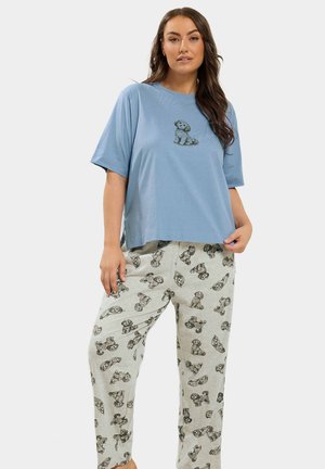 Femme portant un t-shirt oversize bleu clair avec une petite illustration de chien et un pantalon gris clair imprimé avec plusieurs dessins de chiens noirs.