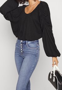 Svart överdimensionerad topp med samlad halsringning och voluminösa ärmar, tillsammans med högmidjade blå jeans med silverknappar som prydnad.