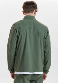 Zielona, lekka kurtka z kołnierzem, elastycznymi mankietami i luźnym krojem. Posiada gładką teksturę oraz minimalne detale szwów na plecach.