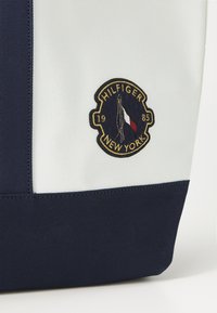 Torbica tamnoplave i bijele boje s okruglim izvezenim logom koji sadrži natpis "HILFIGER NEW YORK" i dizajn zastave. Izdržljiv materijal s kontrastnim teksturama.