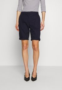 Tmavě modré bavlněné šortky s ohrnutými lemy, na rovný předek a zapínáním na knoflík. Modelka má na sobě černé boty na podpatku se špičatou špičkou a pruhovaný top s dlouhým rukávem.