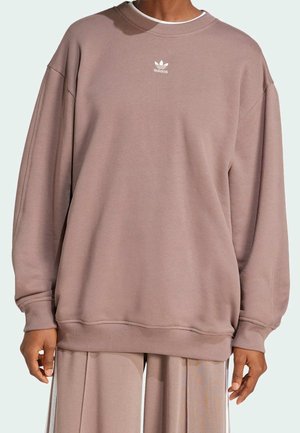 Persona che indossa una felpa Adidas rosa polvere oversize con piccolo logo bianco al centro del petto e pantaloni larghi coordinati.