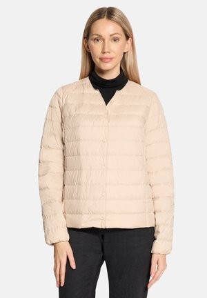 Femme portant une veste matelassée beige clair avec des boutons-pression sur un col roulé noir, associée à un pantalon noir, debout devant un fond uni.