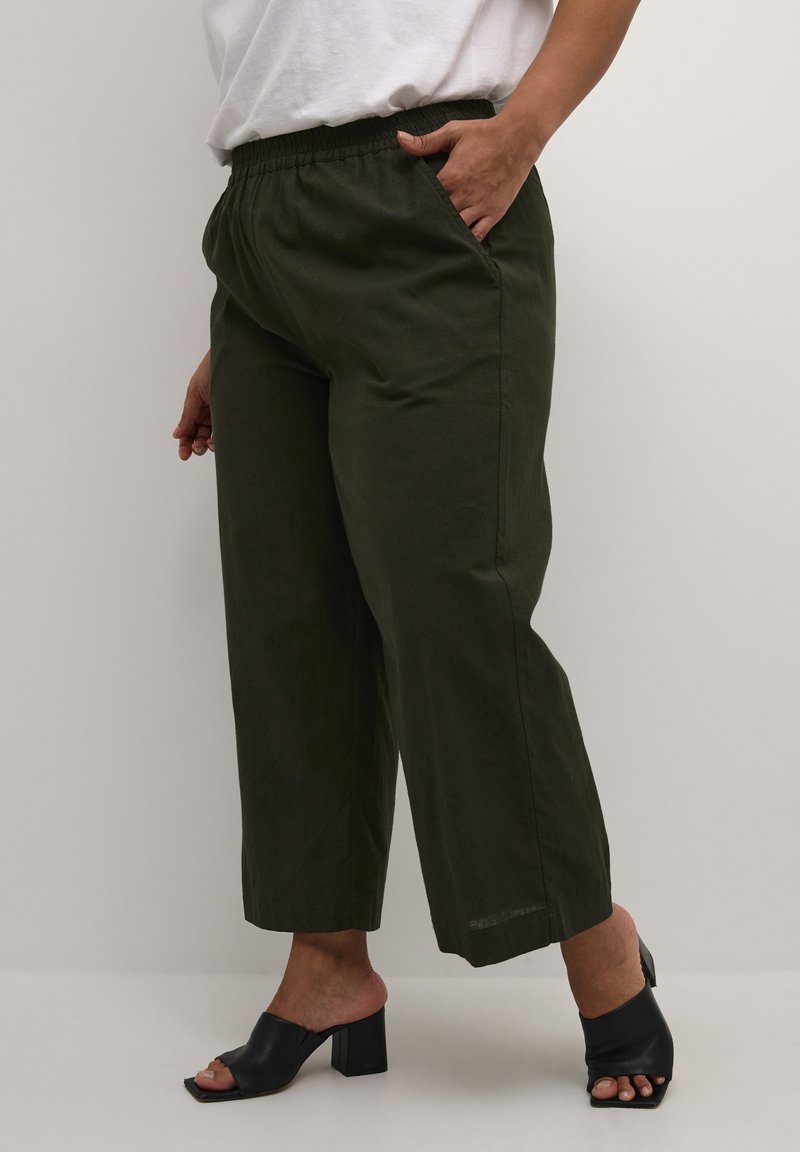 Pantalons évasés vert olive avec une taille élastique et des poches latérales, fabriqués en tissu léger. Associés à des mules noires à bout ouvert.