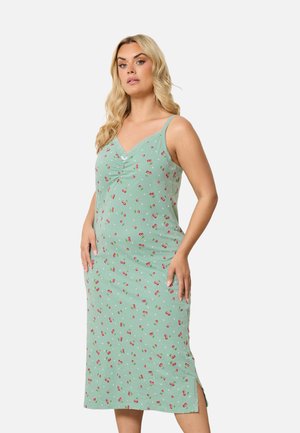 Femme aux longs cheveux blonds portant une robe verte sans manches avec de petits motifs de cerises rouges, debout les mains posées sur les hanches.