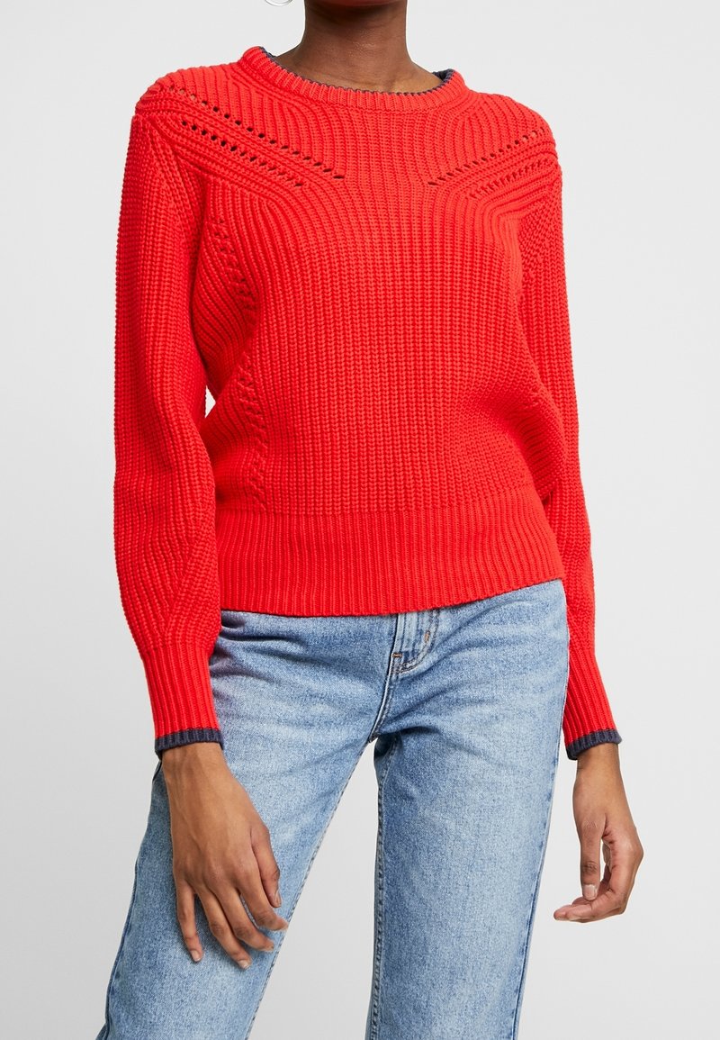 Rød strikket trøje med et tekstureret ribmønster, rund hals og kontrasterende mørke ærmekanter. Kombineret med blå denimjeans.