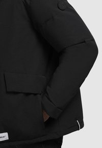 Sort pufferjakke med en blød tekstur, forlommer og et rundt logo på ærmet. Sømløst design med en let oversized pasform.