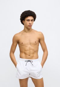 Valged ujumispüksid, millel on elastne vöökoht ja nöör, külgedel peent logokujundust, sile tekstuur ja lõdvestunud lõige.