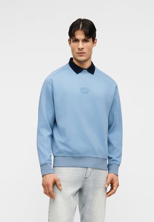 Uomo con felpa azzurro chiaro sopra una camicia con colletto nero e jeans chiari, su sfondo neutro.