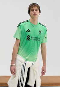 Zelený športový dres s gradientným vzorom, čiernymi akcentmi a logom Liverpool FC. Nosí sa s bielym umelým kožušinovým plášťom a čiernymi nohavicami.