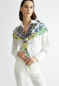 Foulard di seta con stampa leopardata e design floreale in tonalità verdi e blu, annodato attorno al collo, abbinato a una blusa bianca.