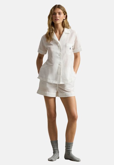 Vrouw staand, gekleed in een wit overhemd met korte mouwen en borstzak, gecombineerd met bijpassende korte broek en grijze gebreide sokken.