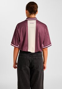 Koszula z krótkim rękawem ma wzór w bordowo-kremowe paski, z kontrastującym, jednolitym tylnym panelem i prążkowanym kołnierzem. Materiał jest gładki.