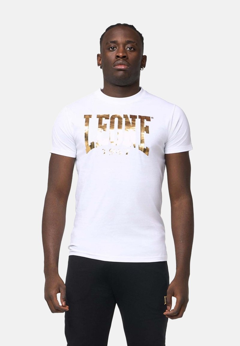 Jeune homme portant un t-shirt blanc avec le logo doré Lonsdale et un pantalon noir, debout devant un fond clair uni.