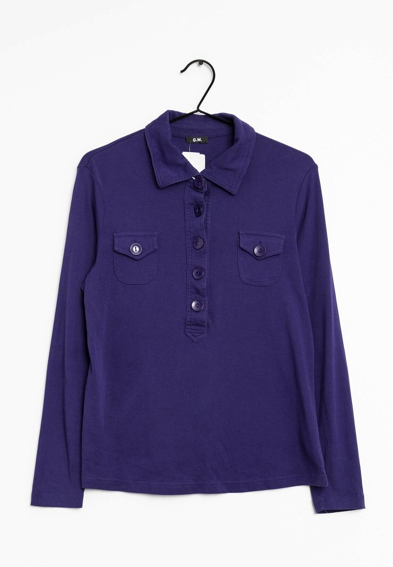 Pitkähihaista violettia polo-paitaa, jossa on kaulus, viisi nappia edessä ja kaksi napilla suljettavaa rintataskua, roikkuu mustassa ripustimessa.