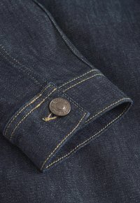 Temno modra denim tkanina z vidno teksturo. Ima srebrno gumb in rumeno šivanje ob robu in šivu.
