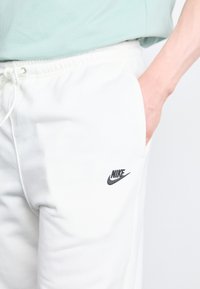 Pantaloni de trening albi din bumbac, cu talie elastică, având un logo negru Nike pe coapsa stângă și buzunare laterale. Textură fină.