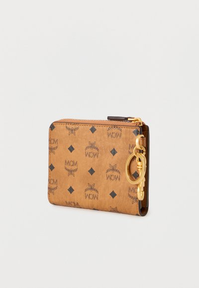 Καφέ δερμάτινο πορτοφόλι με ανάγλυφο λογότυπο MCM, διακοσμητικά μοτίβα σε σχήμα διαμαντιού, χρυστό φερμουάρ και κρίκο για μπρελόκ στο πλάι.