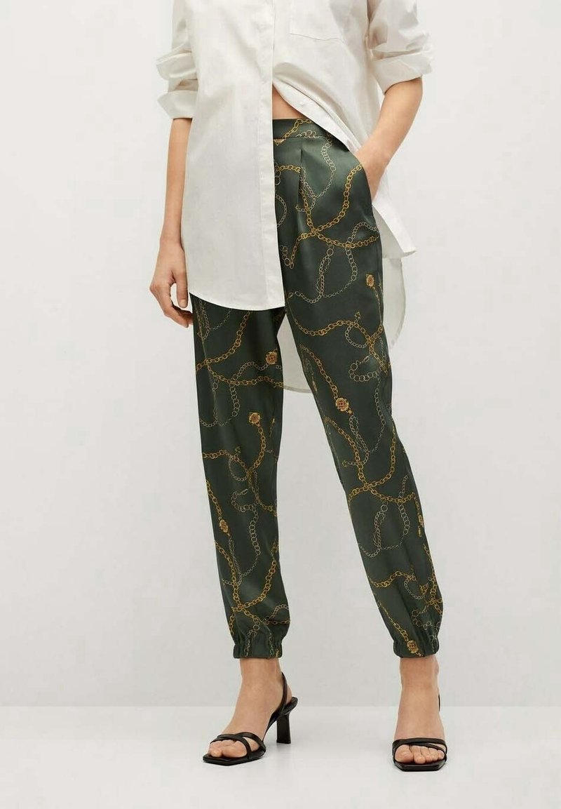 Personne portant des sandales noires à talons, un pantalon vert foncé à imprimé chaîne dorée, et une longue chemise blanche aux manches retroussées.
