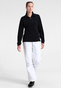 Svart fleece-tröja med dragkedja vid kragen, i kombination med vita byxor med förstärkta knästygn och elastiska ärmslut. Svarta träningsskor bärs.