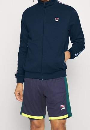 Mężczyzna w granatowej kurtce na zamek i granatowych sportowych szortach z zielonymi i żółtymi panelami po bokach, oba z logo Fila.