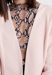 Femme portant une veste rose texturée ouverte sur le devant, par-dessus un haut à col haut avec un imprimé serpent aux tons marron, noir et beige.