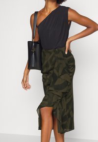 Černý top na jedno rameno v kombinaci s tmavě olivově zelenou sukní s vlnitou texturou a bočním rozparkem. Černá kabelka s detailními třásněmi.