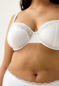 Donna che indossa un reggiseno bianco con pizzo e un perizoma abbinato, mostrando il torace e la parte centrale del corpo con una tonalità e una texture della pelle naturali.