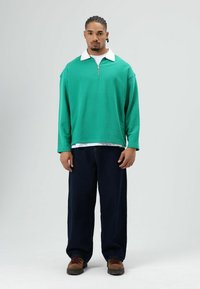 Groene pullover met ritssluiting en witte kraag, losse pasvorm, lange mouwen, gecombineerd met donkerblauwe wijde broekspijpen en bruine schoenen.