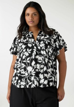 Donna che indossa una blusa nera con stampa floreale bianca e maniche corte arricciate, abbinata a pantaloni neri su uno sfondo semplice.