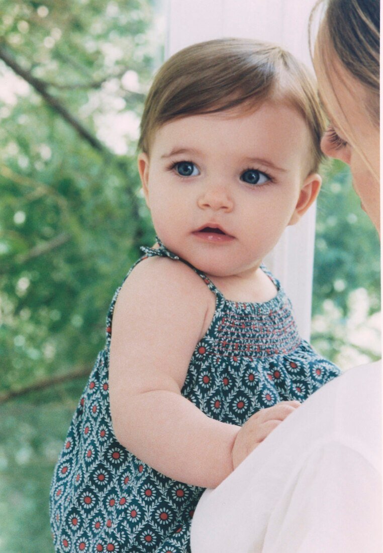 Un tout-petit aux yeux bleus portant une robe sans manches à motifs est tenu par une femme en chemise blanche près d'une fenêtre avec des arbres verts à l'extérieur.
