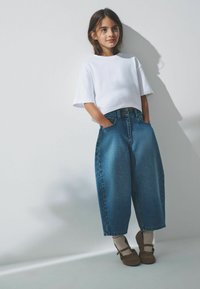 Weißes Crop-Top, blaue hochgeschnittene, lockere Jeans, beige Schuhe mit Knöchelriemen und graue Socken. Einfarbiger Hintergrund mit sanften Schatten.