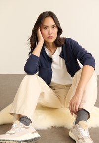 Mörkblå stickad cardigan över en vit t-shirt, enklat med beige vida byxor och ljusgrå New Balance sneakers. Mjuk beige matta under.